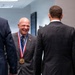 A bald man wearing a gold medal on a red, white, and blue ribbon lanyard is laughing and conversing with two other men in dark suits in what appears to be an official or ceremonial setting. The medal appears to be a distinguished honor or award. Flags, including what appears to be a U.S.-related flag, are visible in the background.