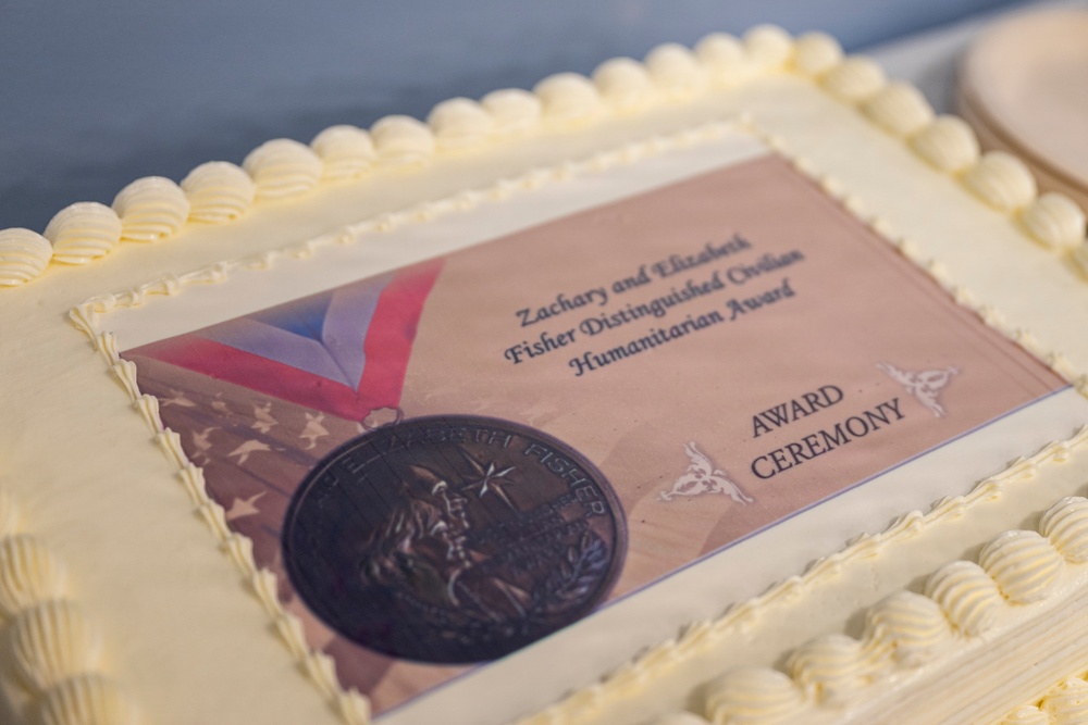 A white frosted sheet cake decorated with an edible printed image commemorating the Zachary and Elizabeth Fisher Distinguished Civilian Humanitarian Award Ceremony. The cake features white buttercream border piping and a printed topper showing the award medal with a red, white, and blue ribbon against an American flag background. This appears to be a celebratory cake prepared for the award ceremony event.