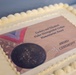 A white frosted sheet cake decorated with an edible printed image commemorating the Zachary and Elizabeth Fisher Distinguished Civilian Humanitarian Award Ceremony. The cake features white buttercream border piping and a printed topper showing the award medal with a red, white, and blue ribbon against an American flag background. This appears to be a celebratory cake prepared for the award ceremony event.