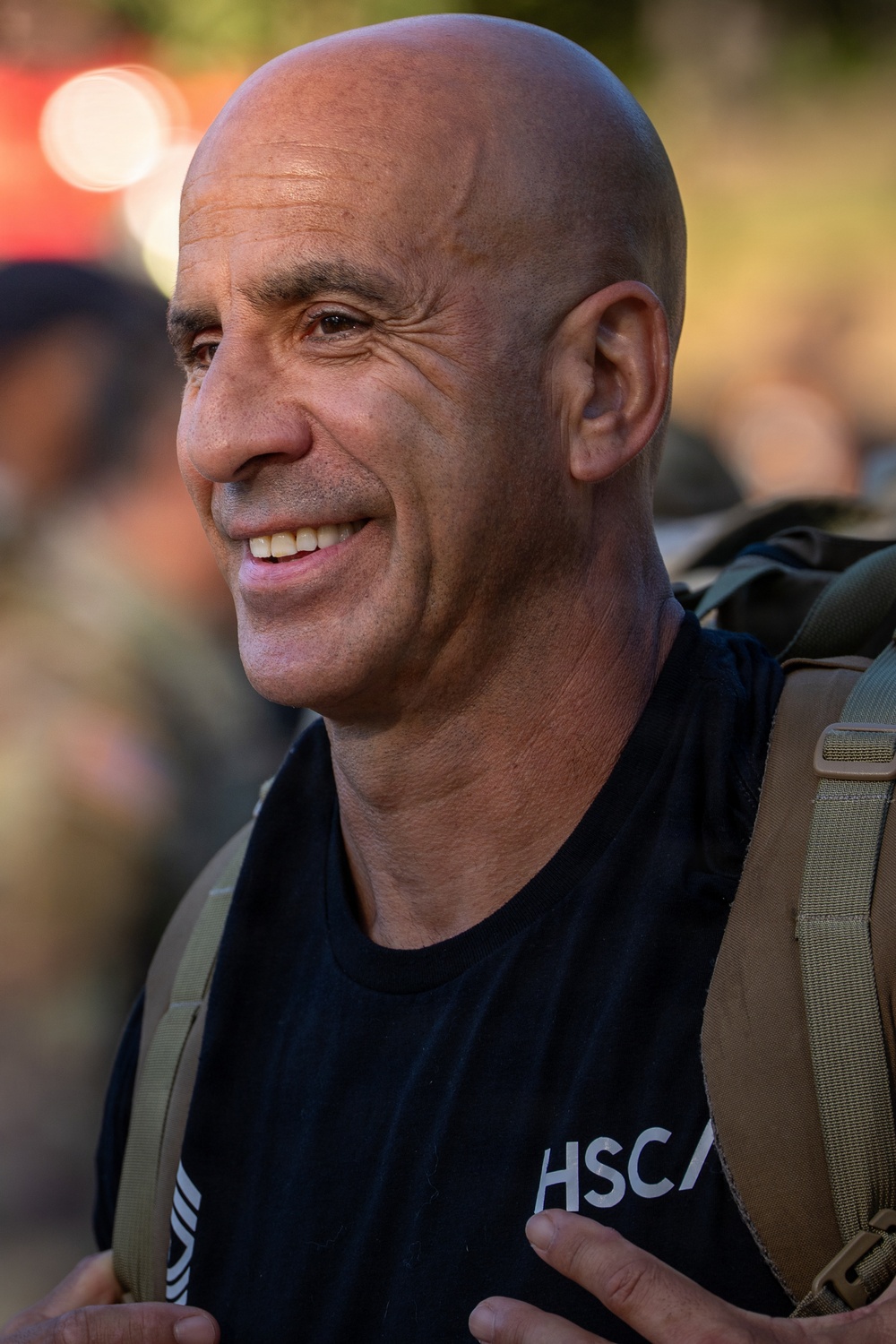A bald middle-aged man is smiling and looking slightly to his left. He is wearing a black t-shirt with partial text reading 'HSCA' and carrying a tactical/military-style backpack. The background is blurred but suggests an outdoor setting with other people present.