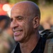 A bald middle-aged man is smiling and looking slightly to his left. He is wearing a black t-shirt with partial text reading 'HSCA' and carrying a tactical/military-style backpack. The background is blurred but suggests an outdoor setting with other people present.