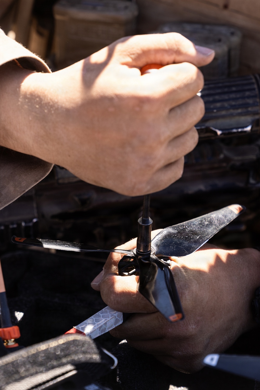 A person is using a screwdriver to work on what appears to be a small drone or RC helicopter propeller assembly in an outdoor setting. Two hands are visible — one gripping the screwdriver handle and the other steadying the propeller hub. The work appears to be taking place near or inside a vehicle, with engine components visible in the background.