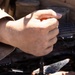 A person is using a screwdriver to work on what appears to be a small drone or RC helicopter propeller assembly in an outdoor setting. Two hands are visible — one gripping the screwdriver handle and the other steadying the propeller hub. The work appears to be taking place near or inside a vehicle, with engine components visible in the background.