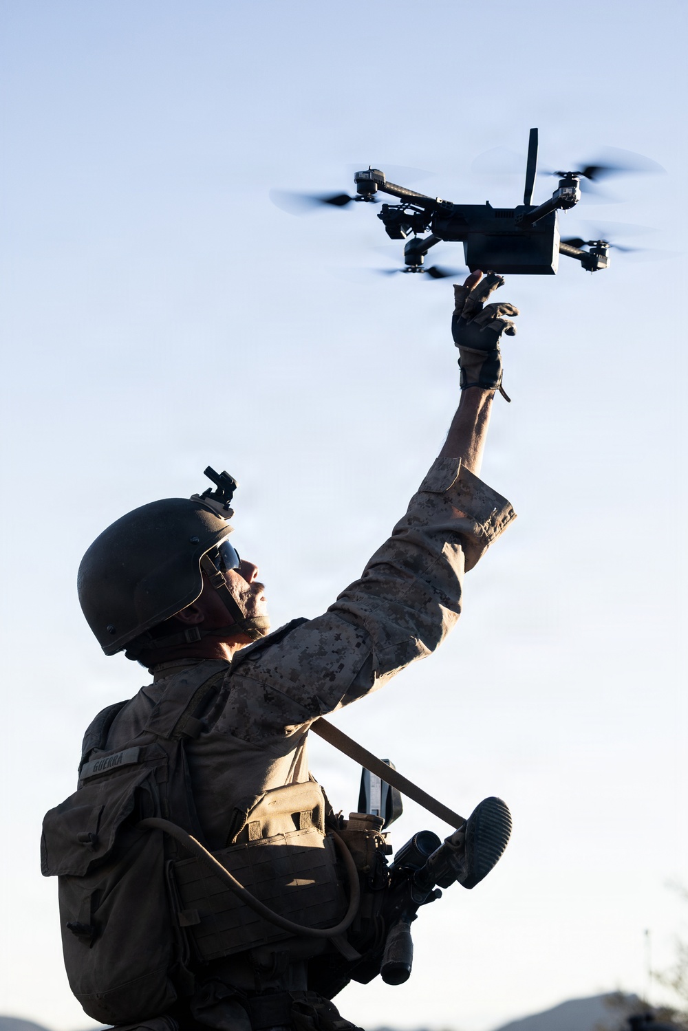 A military service member in full combat gear is launching or recovering a small quadcopter drone by hand. The soldier is wearing MARPAT camouflage uniform, a combat helmet with night vision mount, tactical gloves, and body armor. The image is captured from a low angle against a clear sky, emphasizing the drone deployment action.