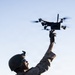 A military service member in full combat gear is launching or recovering a small quadcopter drone by hand. The soldier is wearing MARPAT camouflage uniform, a combat helmet with night vision mount, tactical gloves, and body armor. The image is captured from a low angle against a clear sky, emphasizing the drone deployment action.