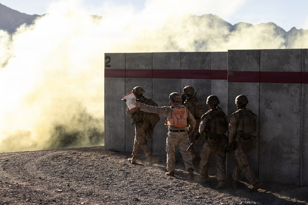 A group of military personnel in desert camouflage uniforms are positioned against a concrete barrier wall marked with a red stripe and the number '2'. One individual wearing an orange safety vest appears to be briefing or directing the others while holding a clipboard or document. A large cloud of smoke or dust is visible to the left, suggesting an active training exercise in a desert or arid environment with mountains visible in the background.