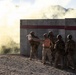 A group of military personnel in desert camouflage uniforms are positioned against a concrete barrier wall marked with a red stripe and the number '2'. One individual wearing an orange safety vest appears to be briefing or directing the others while holding a clipboard or document. A large cloud of smoke or dust is visible to the left, suggesting an active training exercise in a desert or arid environment with mountains visible in the background.