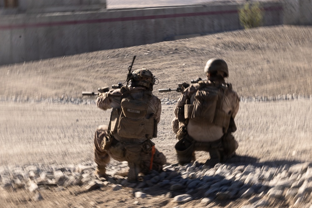 Two armed military personnel in full combat gear are kneeling in a low-profile position on rocky, arid terrain, with weapons raised toward a concrete structure in the background. The image is captured with motion blur applied radially, suggesting dynamic movement or a stylized photographic effect. The environment appears to be a dry, desert-like operational area.