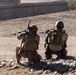 Two armed military personnel in full combat gear are kneeling in a low-profile position on rocky, arid terrain, with weapons raised toward a concrete structure in the background. The image is captured with motion blur applied radially, suggesting dynamic movement or a stylized photographic effect. The environment appears to be a dry, desert-like operational area.