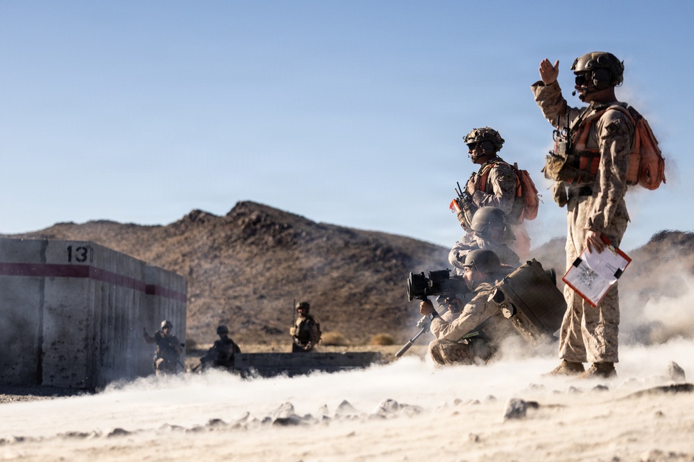 Military personnel in desert camouflage uniforms are conducting what appears to be a training exercise in an arid desert environment. Smoke or dust is visible on the ground, and soldiers are in various tactical positions including prone, kneeling, and standing. A concrete structure marked with the number '13' and a red stripe is visible in the background.
