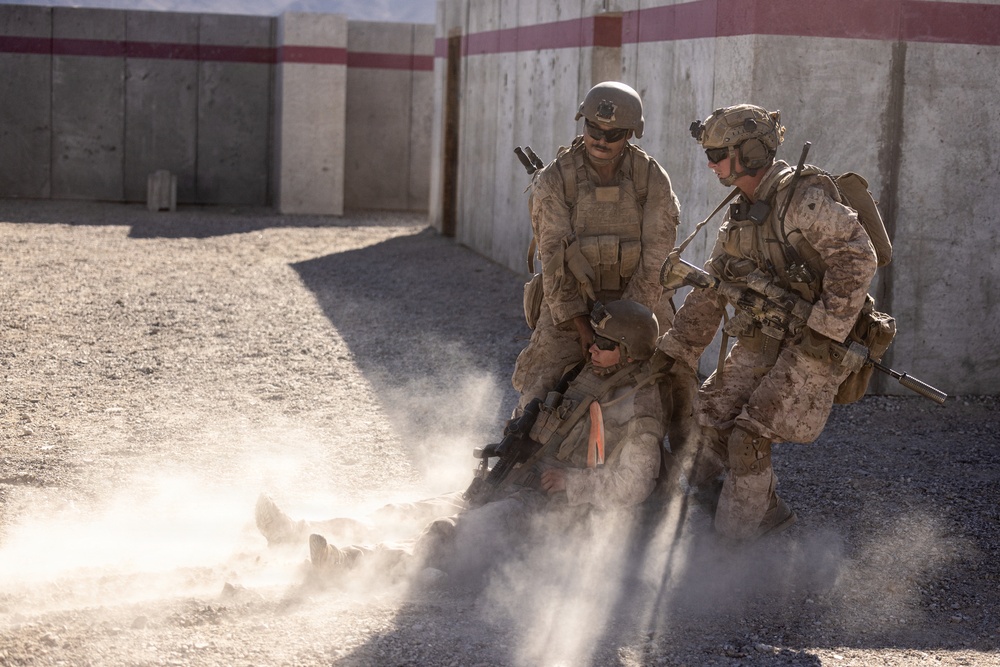 Two standing military personnel in desert camouflage uniforms and tactical gear are assisting or dragging a third downed soldier across a gravel surface, raising a dust cloud. The scene appears to be a casualty evacuation drill or combat training exercise at a military facility. A concrete wall with red and white striping is visible in the background.