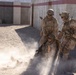 Two standing military personnel in desert camouflage uniforms and tactical gear are assisting or dragging a third downed soldier across a gravel surface, raising a dust cloud. The scene appears to be a casualty evacuation drill or combat training exercise at a military facility. A concrete wall with red and white striping is visible in the background.