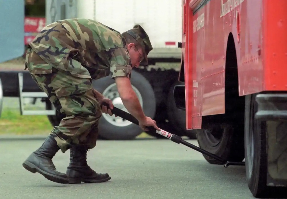 A military personnel wearing woodland camouflage BDUs and a garrison cap is bent over inspecting the underside of a large red vehicle, likely a truck or fire apparatus. The soldier is using an extendable mirror or inspection tool to examine beneath the vehicle, suggesting a security checkpoint or vehicle inspection procedure. The setting appears to be a road or checkpoint area with other vehicles visible in the background.