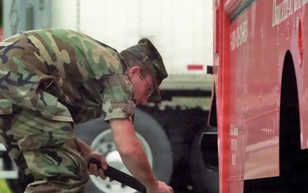 A military personnel wearing woodland camouflage BDUs and a garrison cap is bent over inspecting the underside of a large red vehicle, likely a truck or fire apparatus. The soldier is using an extendable mirror or inspection tool to examine beneath the vehicle, suggesting a security checkpoint or vehicle inspection procedure. The setting appears to be a road or checkpoint area with other vehicles visible in the background.