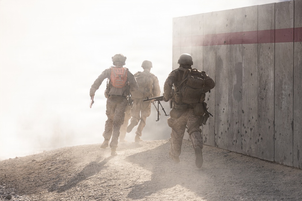 Three military personnel in combat gear are running through a dusty, hazy environment toward a concrete barrier wall marked with a red horizontal stripe. The scene appears to depict a tactical training exercise or operational movement, with significant dust obscuring visibility. The soldiers are moving in formation, carrying weapons and tactical equipment.