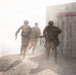 Three military personnel in combat gear are running through a dusty, hazy environment toward a concrete barrier wall marked with a red horizontal stripe. The scene appears to depict a tactical training exercise or operational movement, with significant dust obscuring visibility. The soldiers are moving in formation, carrying weapons and tactical equipment.