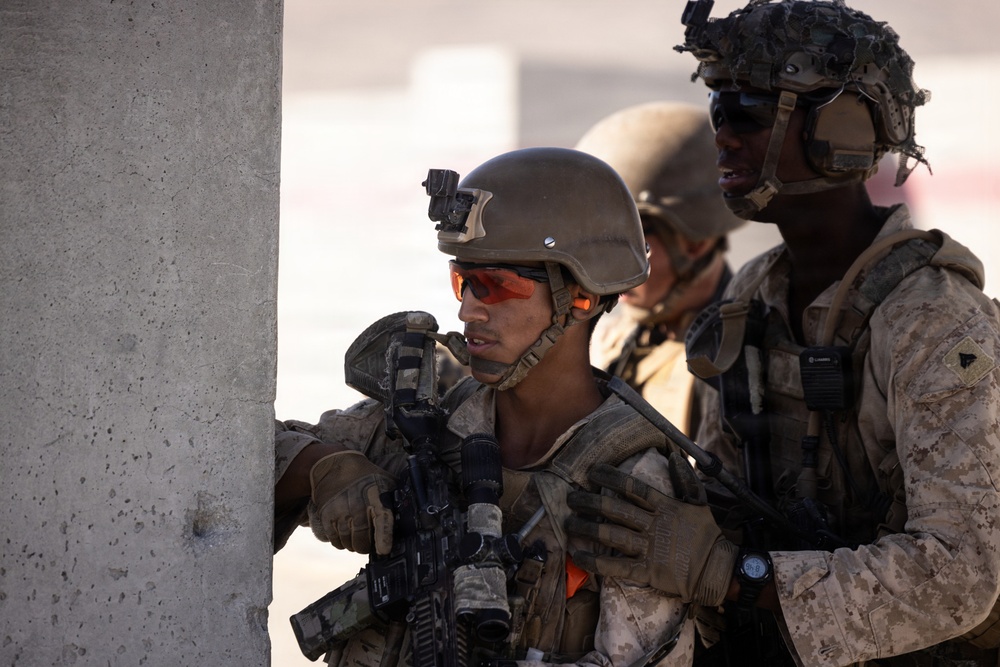 The image shows multiple military personnel in tactical gear taking cover behind a concrete wall or pillar. The soldiers are equipped with helmets, body armor, weapons, and communication devices, suggesting a tactical operation or training exercise. The setting appears to be an urban or built environment in a arid/desert region based on the lighting and background.