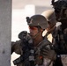 The image shows multiple military personnel in tactical gear taking cover behind a concrete wall or pillar. The soldiers are equipped with helmets, body armor, weapons, and communication devices, suggesting a tactical operation or training exercise. The setting appears to be an urban or built environment in a arid/desert region based on the lighting and background.