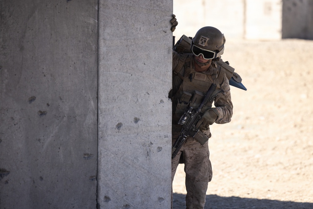 A U.S. military service member in full combat gear is taking cover behind a concrete wall, appearing to conduct urban warfare training or an operational exercise. The soldier is wearing MARPAT camouflage, a combat helmet with goggles, and is armed with an assault rifle. The arid, sandy environment with concrete block structures in the background suggests a training facility or forward operating base.