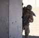 A U.S. military service member in full combat gear is taking cover behind a concrete wall, appearing to conduct urban warfare training or an operational exercise. The soldier is wearing MARPAT camouflage, a combat helmet with goggles, and is armed with an assault rifle. The arid, sandy environment with concrete block structures in the background suggests a training facility or forward operating base.