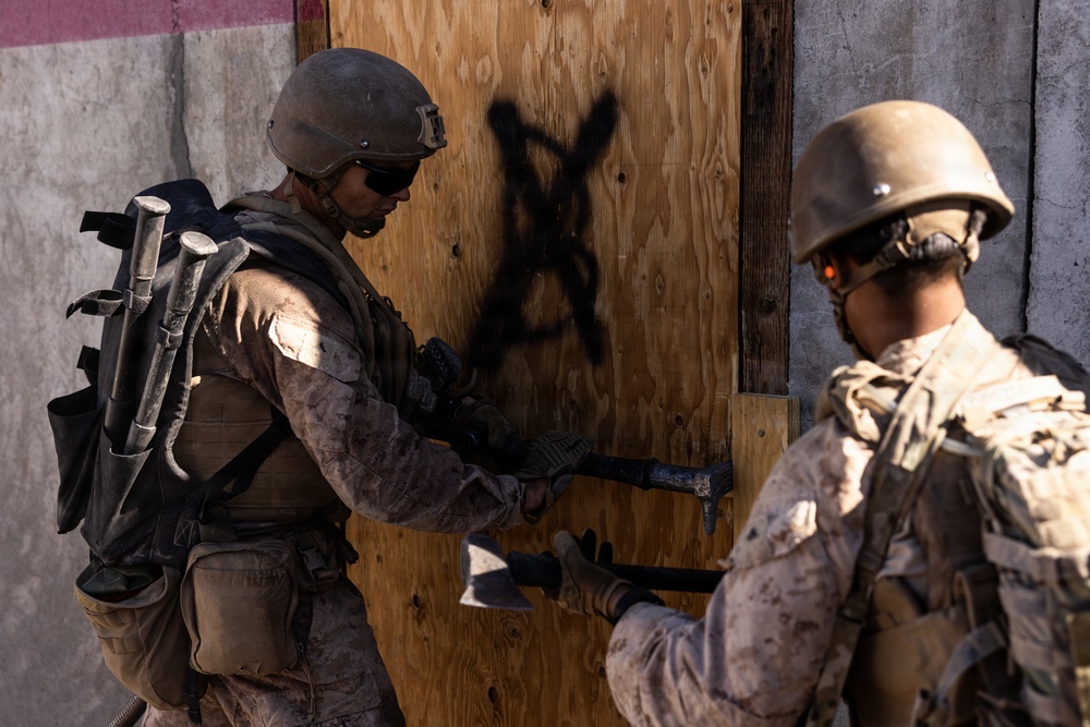 Two military personnel in desert camouflage uniforms are conducting a breaching operation on a wooden door marked with a black spray-painted X. The soldier in the foreground is using a breaching tool (Halligan bar or similar pry tool) to force open the door, while a second soldier stands nearby in support. This appears to be a training exercise based on the controlled environment and mock structure.