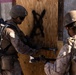 Two military personnel in desert camouflage uniforms are conducting a breaching operation on a wooden door marked with a black spray-painted X. The soldier in the foreground is using a breaching tool (Halligan bar or similar pry tool) to force open the door, while a second soldier stands nearby in support. This appears to be a training exercise based on the controlled environment and mock structure.