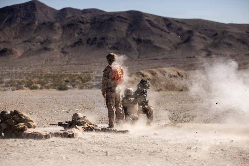 Military personnel in desert camouflage uniforms are conducting a live-fire training exercise in an arid desert environment. A weapon system, likely a rocket launcher or recoilless rifle, has just been fired, producing a visible backblast cloud of smoke and debris. One standing soldier wearing an orange safety vest/pack appears to be an observer or safety officer overseeing the exercise.