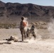 Military personnel in desert camouflage uniforms are conducting a live-fire training exercise in an arid desert environment. A weapon system, likely a rocket launcher or recoilless rifle, has just been fired, producing a visible backblast cloud of smoke and debris. One standing soldier wearing an orange safety vest/pack appears to be an observer or safety officer overseeing the exercise.