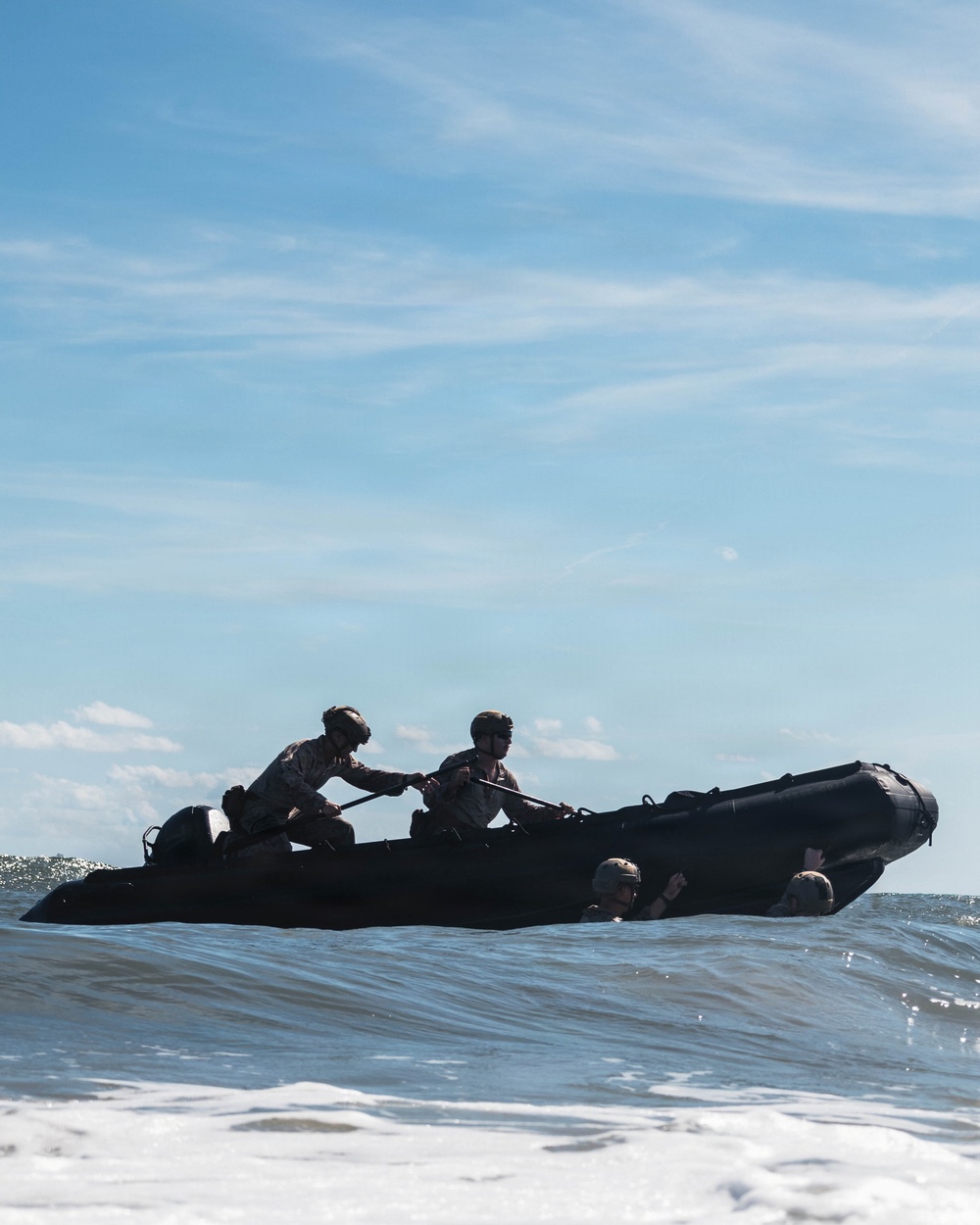 Military personnel in camouflage uniforms and helmets are operating a black inflatable rubber raiding craft (IBS/Zodiac-type boat) in ocean surf conditions. At least four individuals are visible, with two actively paddling at the bow and others positioned lower in or alongside the craft. This appears to be an amphibious training exercise, consistent with Marine Corps or special operations beach landing drills.