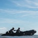 Military personnel in camouflage uniforms and helmets are operating a black inflatable rubber raiding craft (IBS/Zodiac-type boat) in ocean surf conditions. At least four individuals are visible, with two actively paddling at the bow and others positioned lower in or alongside the craft. This appears to be an amphibious training exercise, consistent with Marine Corps or special operations beach landing drills.