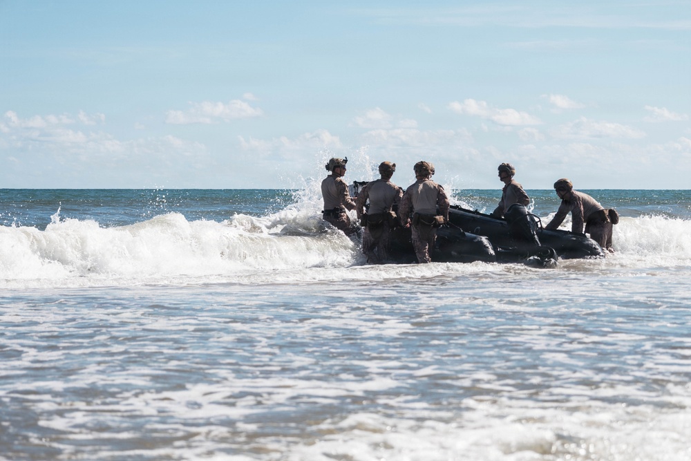 A group of military personnel in combat gear and helmets are maneuvering a black inflatable rubber raiding craft (IBS/Zodiac-type boat) through breaking surf at a beach. The soldiers appear to be conducting an amphibious beach landing or surf passage training exercise. Waves are crashing against the boat and personnel as they push through the surf zone.