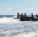 A group of military personnel in combat gear and helmets are maneuvering a black inflatable rubber raiding craft (IBS/Zodiac-type boat) through breaking surf at a beach. The soldiers appear to be conducting an amphibious beach landing or surf passage training exercise. Waves are crashing against the boat and personnel as they push through the surf zone.