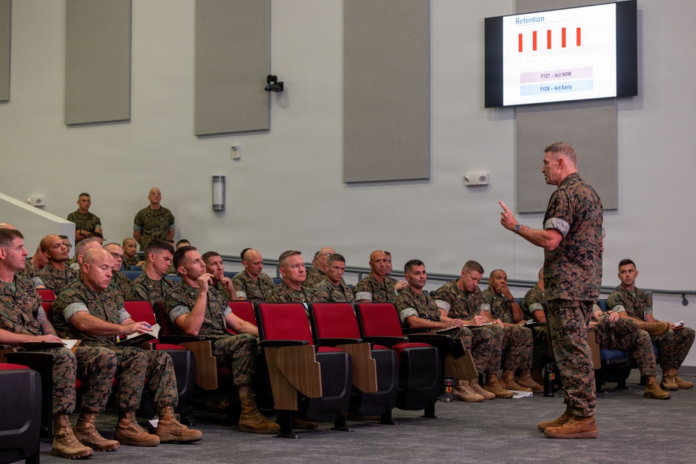 A military briefing is taking place in a modern auditorium where a senior military officer in camouflage uniform addresses a seated audience of fellow service members. The presenter is gesturing toward a projected screen displaying a 'Retention' bar chart with fiscal year data labeled 'FY27 - Act NOW' and 'FY28 - Act Early'. The audience, all wearing matching camouflage uniforms consistent with U.S. Marine Corps MARPAT pattern, are attentively listening and some are taking notes.