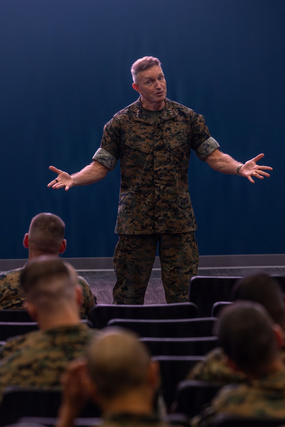 A senior U.S. Marine Corps officer in MARPAT digital camouflage uniform is addressing an audience of Marines in what appears to be a briefing room or auditorium. The speaker has general officer rank insignia visible on his collar and is gesturing with both arms outstretched, suggesting an engaged, emphatic presentation. Multiple Marines in matching camouflage uniforms are seated in the audience facing the speaker.