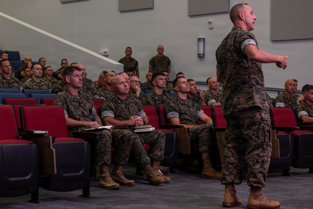 A military briefing or lecture is taking place in a modern auditorium-style room. A standing service member in camouflage uniform appears to be addressing a seated audience of fellow military personnel, gesturing with his right arm. The audience members are attentively listening, with some taking notes.