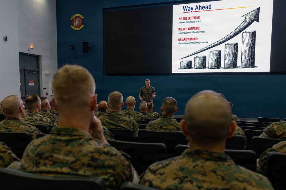 A military briefing is taking place in an auditorium-style room where a uniformed presenter addresses a seated audience of Marines in camouflage uniforms. The presenter stands in front of a large projection screen displaying a slide titled 'Way Ahead' with a bar chart and three key points about listening, adapting, and winning. A United States Marine Corps emblem is visible on the teal-colored wall.