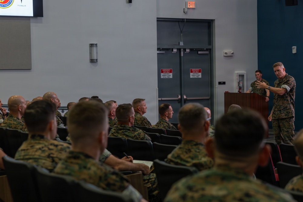 A military briefing or lecture is taking place in an auditorium-style room where a speaker in camouflage uniform addresses a seated audience of fellow service members. The speaker stands at a wooden podium on the right side of the room, gesturing with both hands while delivering remarks. The audience, all wearing matching camouflage uniforms consistent with U.S. Marine Corps MARPAT pattern, are seated in rows of chairs facing the presenter.
