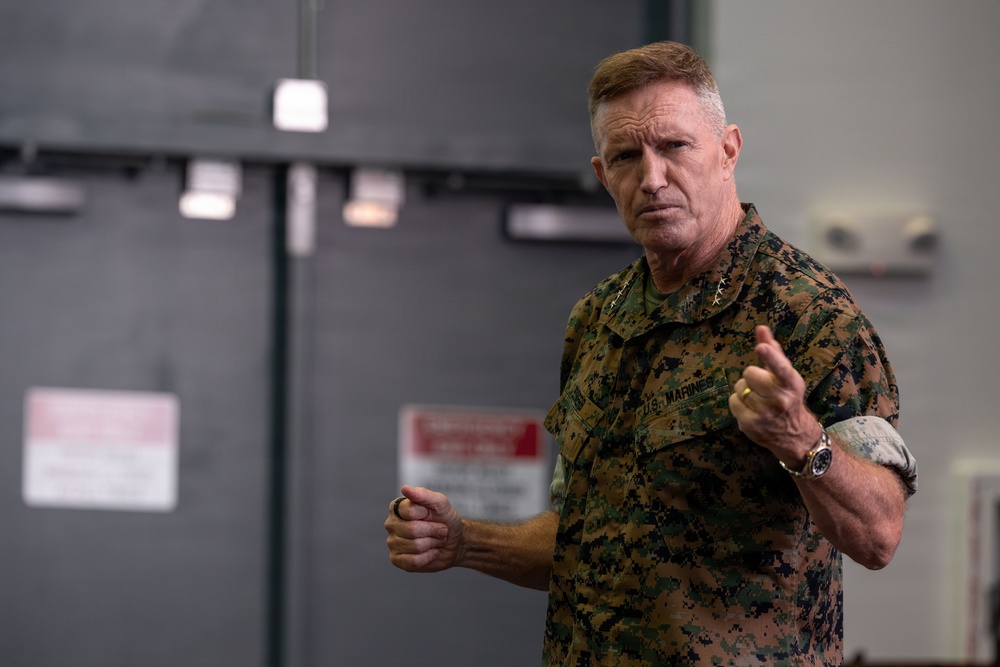 A senior U.S. Marine Corps officer in MARPAT digital camouflage uniform is photographed in what appears to be an indoor military facility. He is gesturing with both hands in a manner suggesting he is speaking or giving a briefing. His collar insignia indicates a high-ranking general officer.