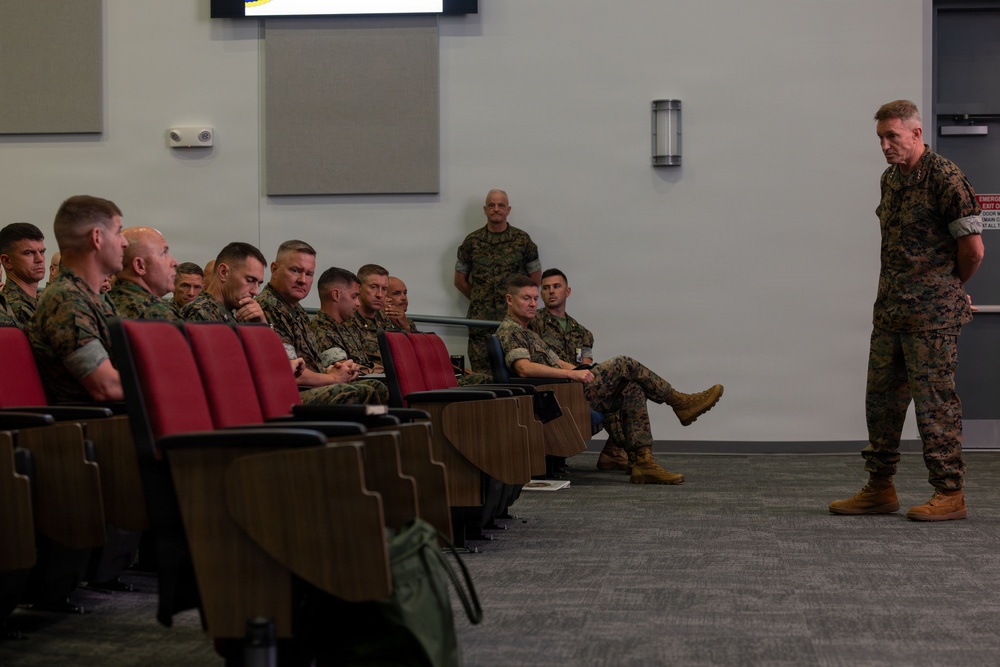 A military briefing or address is taking place in a modern auditorium-style room. A senior military officer in camouflage uniform stands at the front addressing a seated group of uniformed personnel, likely U.S. Marines based on the MARPAT camouflage pattern. The setting appears to be a formal military conference or training facility.