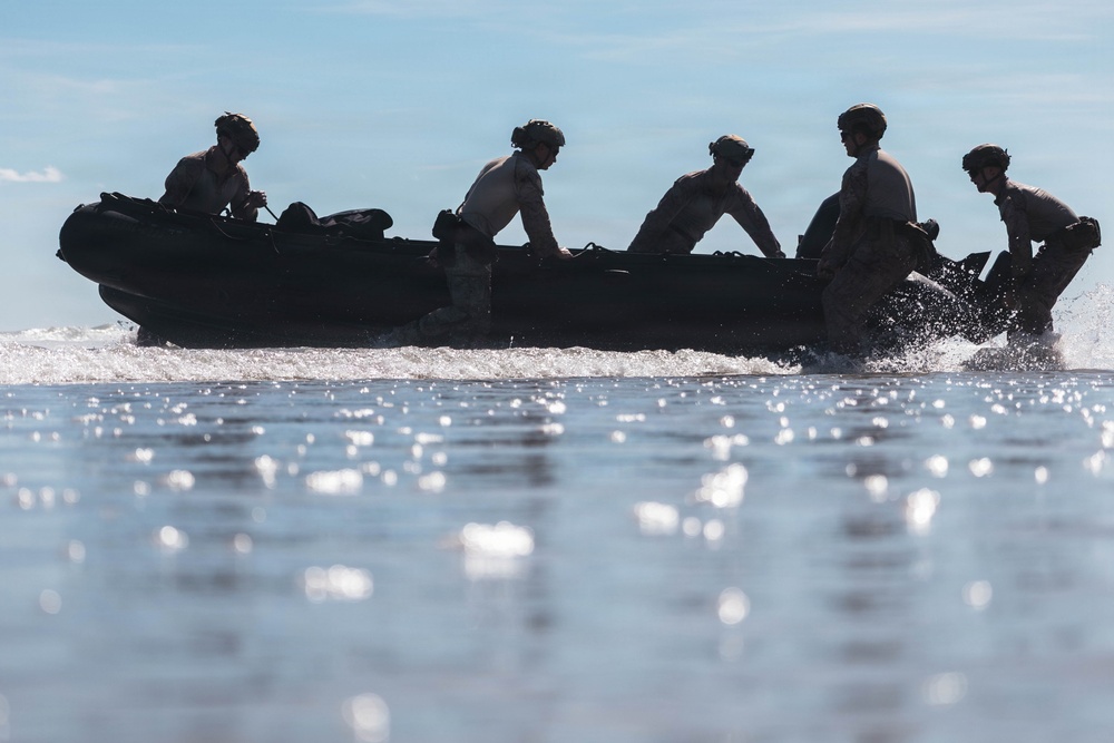 A silhouetted group of approximately five individuals wearing helmets are aboard a dark inflatable rubber boat moving through shallow coastal water. The image is captured in strong backlight, creating a dramatic silhouette effect against a bright sky and reflective water surface. The scene appears to depict a military or tactical waterborne operation.