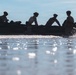 A silhouetted group of approximately five individuals wearing helmets are aboard a dark inflatable rubber boat moving through shallow coastal water. The image is captured in strong backlight, creating a dramatic silhouette effect against a bright sky and reflective water surface. The scene appears to depict a military or tactical waterborne operation.