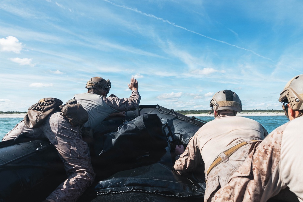 Military personnel in combat/tactical gear are aboard a black inflatable rubber raiding craft (CRRC) approaching a sandy beach shoreline. One individual is raising their hand, likely giving a hand signal to other team members. The personnel are wearing camouflage uniforms and tactical helmets consistent with Marine Corps or special operations forces.