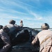 Military personnel in combat/tactical gear are aboard a black inflatable rubber raiding craft (CRRC) approaching a sandy beach shoreline. One individual is raising their hand, likely giving a hand signal to other team members. The personnel are wearing camouflage uniforms and tactical helmets consistent with Marine Corps or special operations forces.
