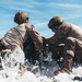 Two military personnel in tactical gear are positioned on what appears to be an inflatable rubber boat (IRB) being hit by ocean waves. Both individuals are wearing camouflage uniforms and tactical helmets, suggesting a maritime amphibious operation or training exercise. The image captures a dynamic moment with significant water spray surrounding the vessel.