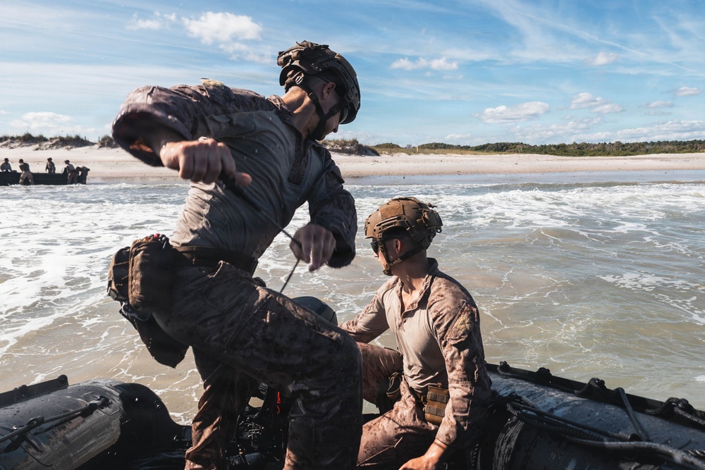 Military personnel in tactical gear are conducting an amphibious beach landing exercise using an inflatable rubber combat boat (CRRC). Two soldiers in the foreground are actively working with the vessel at the shoreline, with one appearing soaked and mud-covered. Additional personnel and boats are visible in the background along the sandy beach.