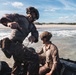 Military personnel in tactical gear are conducting an amphibious beach landing exercise using an inflatable rubber combat boat (CRRC). Two soldiers in the foreground are actively working with the vessel at the shoreline, with one appearing soaked and mud-covered. Additional personnel and boats are visible in the background along the sandy beach.