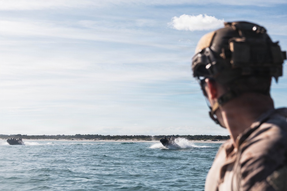 A military personnel wearing a tactical helmet with mounted equipment observes two small inflatable assault craft (likely CRRCs - Combat Rubber Raiding Craft) moving at speed across open water toward a sandy shoreline. The scene appears to be a maritime military exercise or amphibious operation. The foreground subject is out of focus while the watercraft in the middle distance are the focal point of the activity.