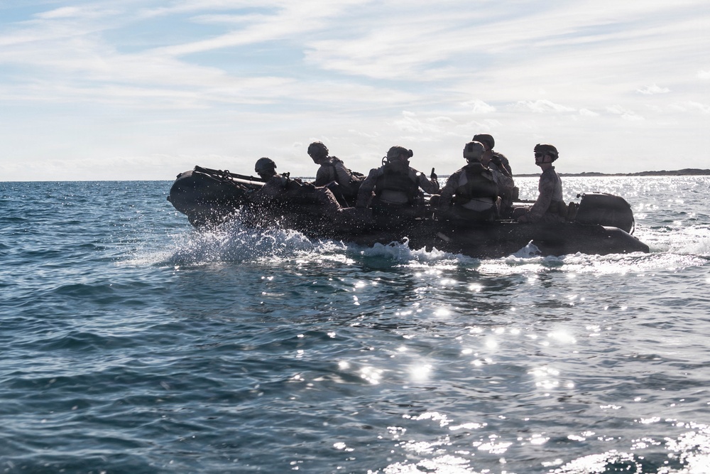 A group of military personnel in full tactical gear are traveling across open water in a dark inflatable rubber assault craft (CRRC). The figures are silhouetted against a bright, sun-reflecting sea surface with a coastline visible in the background. The image captures what appears to be a maritime insertion or amphibious training operation.