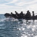 A group of military personnel in full tactical gear are traveling across open water in a dark inflatable rubber assault craft (CRRC). The figures are silhouetted against a bright, sun-reflecting sea surface with a coastline visible in the background. The image captures what appears to be a maritime insertion or amphibious training operation.