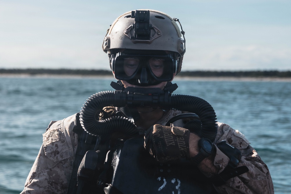 A military combat diver is photographed in close-up wearing full tactical diving gear, including a rebreather mask with corrugated hoses, a tan ballistic helmet with a night-vision mount, and desert camouflage uniform. The subject appears to be in or near open water, with a coastline visible in the background. The image captures a special operations or combat diving context based on the specialized equipment visible.